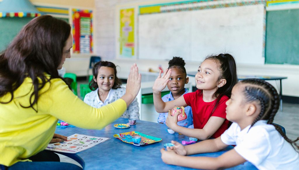 teacher at a small table in a classroom high fiving a young girl
