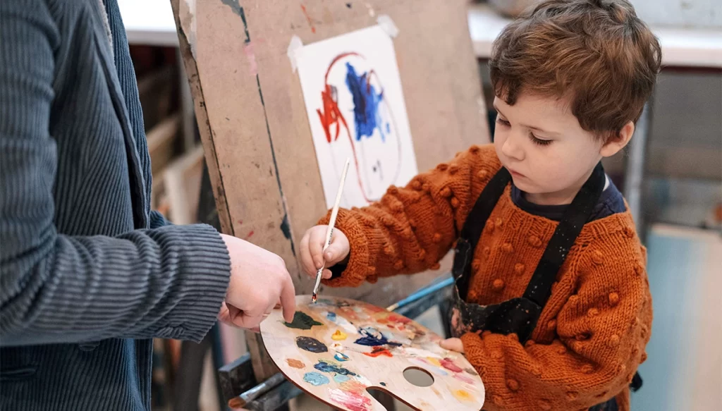 young boy mixing paint on a pallette while the teacher points at the pallette