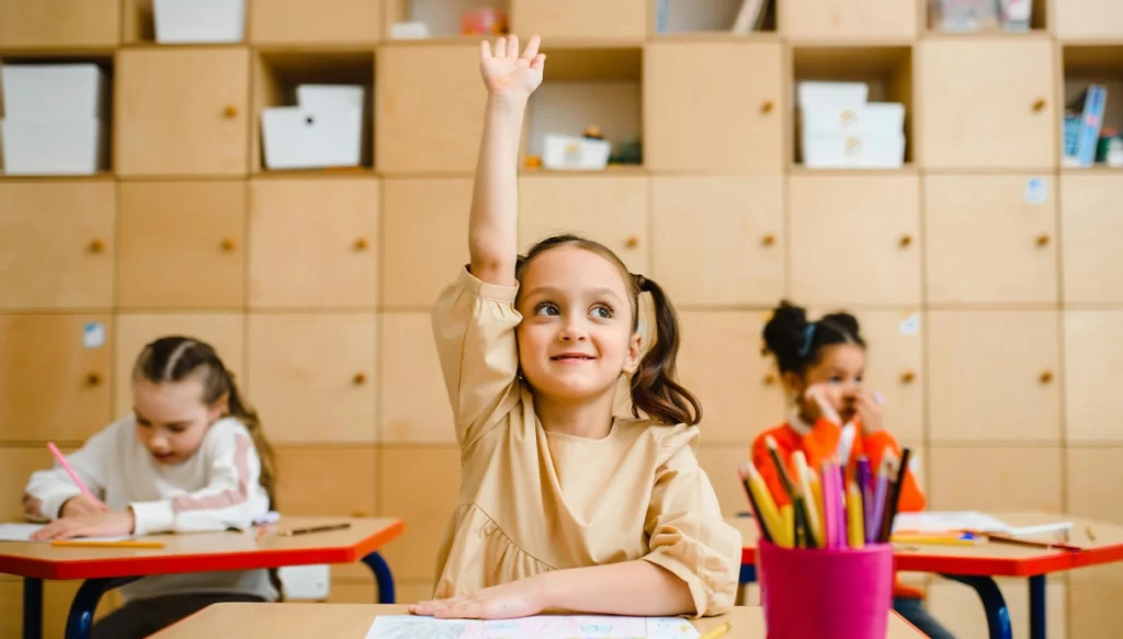 young girl at her deak in a classroom with hand raised