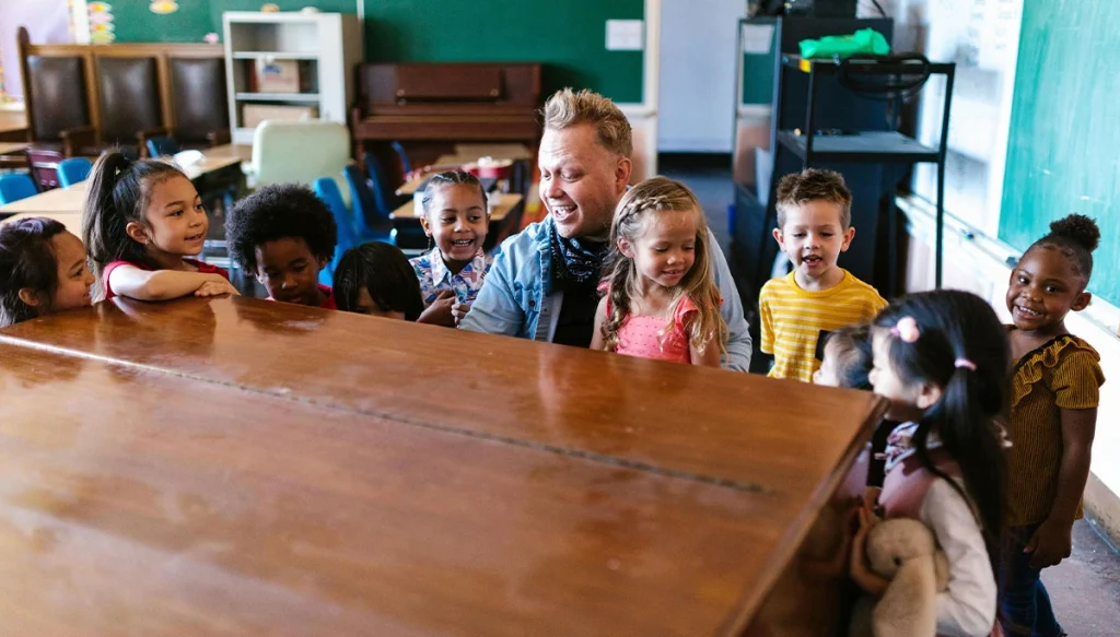 adult male teacher sitting at a piano surrounded by other young students